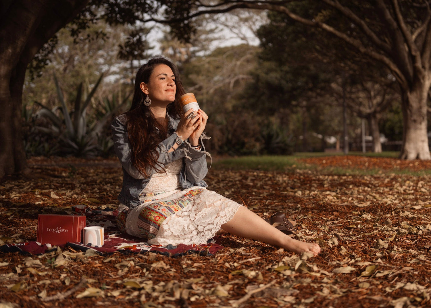 Woman sitting on a blanket in a park, holding a tea can, with a scenic background.