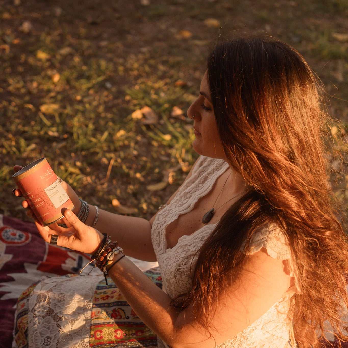 Woman sitting on a blanket outdoors, holding a pink tea tin.
